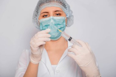 Beyaz önlüklü, tıbbi şapkalı, maskeli ve eldivenli kadın doktor şırınga iğnesi ve tıbbi ampulle çalışıyor. Steril giysiler içindeki bir bilim adamı beyaz arka planda duran bir aşı hazırlıyor. Coronavirus aşısı arama konsepti.