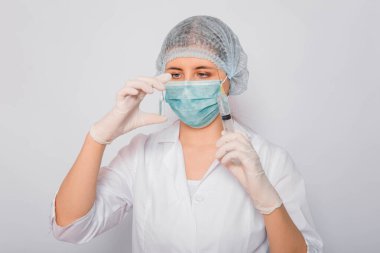 Doktor elinde şırınga ve aşılı bir ampul tutuyor. Beyaz önlüklü, tıbbi maskeli, beyaz arka planda şapka ve eldivenli genç bir kadın. Laboratuvar tıbbi çalışanı. Coronavirus aşısı arama konsepti.