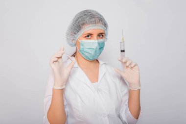 Doktor elinde şırınga ve aşılı bir ampul tutuyor. Beyaz önlüklü, tıbbi maskeli, beyaz arka planda şapka ve eldivenli genç bir kadın. Laboratuvar tıbbi çalışanı. Coronavirus aşısı arama konsepti.