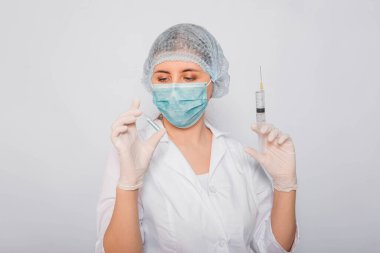 Doktor elinde şırınga ve aşılı bir ampul tutuyor. Beyaz önlüklü, tıbbi maskeli, beyaz arka planda şapka ve eldivenli genç bir kadın. Laboratuvar tıbbi çalışanı. Coronavirus aşısı arama konsepti.