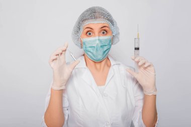 Doktor elinde şırınga ve aşılı bir ampul tutuyor. Beyaz önlüklü, tıbbi maskeli, beyaz arka planda şapka ve eldivenli genç bir kadın. Laboratuvar tıbbi çalışanı. Coronavirus aşısı arama konsepti.