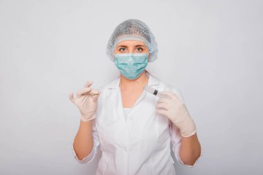 Doktor elinde şırınga ve aşılı bir ampul tutuyor. Beyaz önlüklü, tıbbi maskeli, beyaz arka planda şapka ve eldivenli genç bir kadın. Laboratuvar tıbbi çalışanı. Coronavirus aşısı arama konsepti.