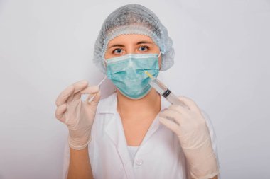Doktor elinde şırınga ve aşılı bir ampul tutuyor. Beyaz önlüklü, tıbbi maskeli, beyaz arka planda şapka ve eldivenli genç bir kadın. Laboratuvar tıbbi çalışanı. Coronavirus aşısı arama konsepti.