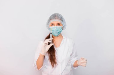 Beyaz önlüklü, maskeli, şapkalı ve eldivenli kadın bir şırıngayı elinde tutuyor. Beyaz arka planda duruyor. Coronavirus aşısı arama konsepti.