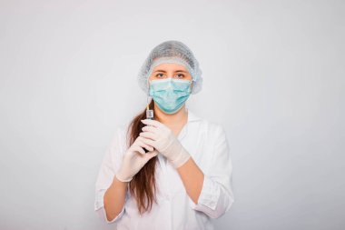 Beyaz önlüklü, maskeli, şapkalı ve eldivenli kadın bir şırıngayı elinde tutuyor. Beyaz arka planda duruyor. Coronavirus aşısı arama konsepti.