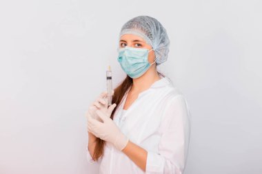 Beyaz önlüklü, maskeli, şapkalı ve eldivenli kadın bir şırıngayı elinde tutuyor. Beyaz arka planda duruyor. Coronavirus aşısı arama konsepti.