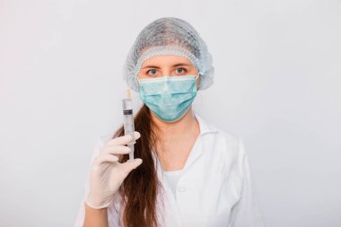 Beyaz önlüklü, maskeli, şapkalı ve eldivenli kadın bir şırıngayı elinde tutuyor. Beyaz arka planda duruyor. Coronavirus aşısı arama konsepti.