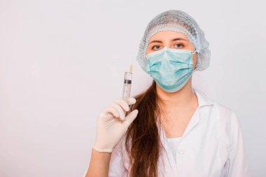 Beyaz önlüklü, maskeli, şapkalı ve eldivenli kadın bir şırıngayı elinde tutuyor. Beyaz arka planda duruyor. Coronavirus aşısı arama konsepti.