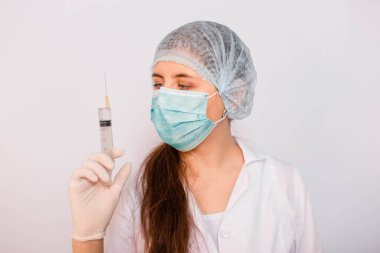 Beyaz önlüklü, maskeli, şapkalı ve eldivenli kadın bir şırıngayı elinde tutuyor. Beyaz arka planda duruyor. Coronavirus aşısı arama konsepti.
