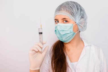 Beyaz önlüklü, maskeli, şapkalı ve eldivenli kadın bir şırıngayı elinde tutuyor. Beyaz arka planda duruyor. Coronavirus aşısı arama konsepti.