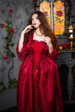 Beautiful girl in a magnificent, red rococo dress. Against the background of the fireplace, window, and flowers. clipart