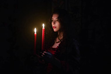 Muhteşem kırmızı rokoko elbiseli güzel bir kız elinde mumlu bir şamdan tutuyor, karanlıkta duruyor, şöminenin, pencerenin ve çiçeklerin arkasında..