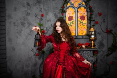 A beautiful girl in a magnificent red dress of the Rococo era stands against a fireplace, a window and flowers with a lamp with candles in her hands. clipart