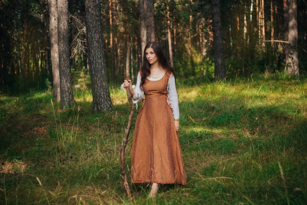 中世のカウボーイ服の若い美しい少女は 手に棒で 地面に裸足だ 森と緑の草を背景に きれいな肌をしたモデル — ストック写真