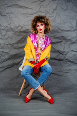 Sarı ceketli, mavi kotlu, afro saçlı bir kız sandalyede oturuyor. Moda seksenleri, disko çağı. Gri arkaplanda stüdyo fotoğrafı.