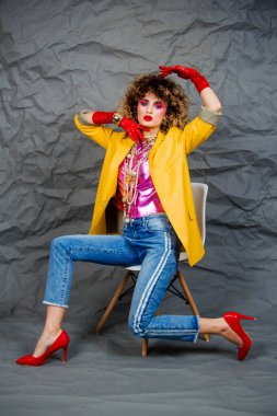 Sarı ceketli, mavi kotlu, afro saçlı bir kız sandalyede oturuyor. Moda seksenleri, disko çağı. Gri arkaplanda stüdyo fotoğrafı.