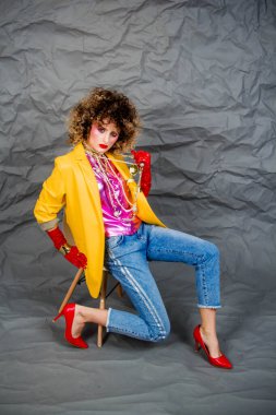 Sarı ceketli, mavi kotlu, afro saçlı bir kız sandalyede oturuyor. Moda seksenleri, disko çağı. Gri arkaplanda stüdyo fotoğrafı.