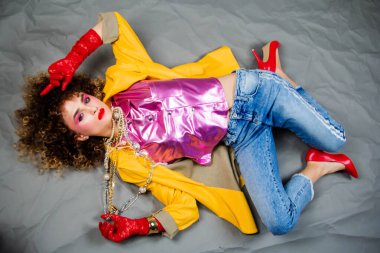 Sarı ceketli ve saçı afro saçlı mavi kotlu kız yerde yatıyor. Moda seksenleri, disko çağı. Gri arkaplanda stüdyo fotoğrafı.
