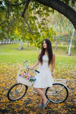 Beyaz etekli, bisikletli ve çiçek sepetli bir kız, sonbaharda, akşamları, parkta sarı yaprakların üzerinde duruyor..