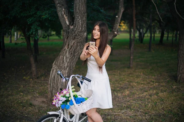 白いスカートの女の子とスマートフォンを手にしたベストは 自転車や公園の花のバスケットの横に立っています — ストック写真