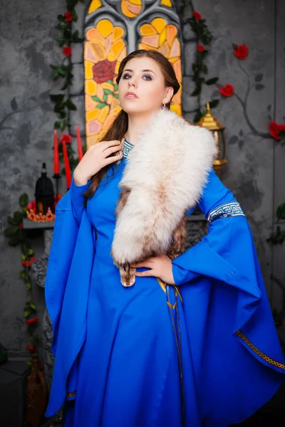 Young beautiful girl in a medieval blue dress with long sleeves and fur on her shoulders posing hands clasped together. Photo on the background of a window, fireplace and flowers.