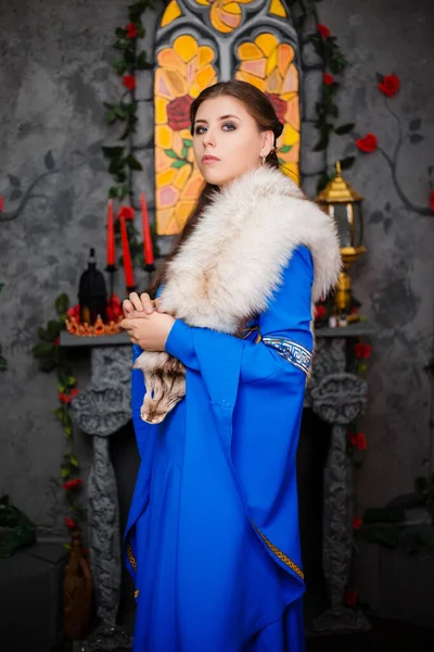 穿着中世纪蓝色连衣裙 头戴长袖 肩上披着毛皮的年轻漂亮姑娘双手紧握在一起 照片在窗户 壁炉和花朵的背景上 — 图库照片