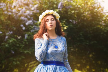 Mavili muhteşem elbiseli çekici bir kadın çiçek açan leylak çiçeklerinin arka planında parkta yürüyor..