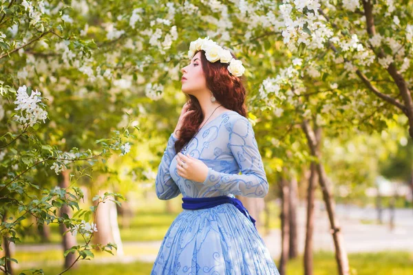 頭に白い花の縁がある青いパフィーのドレスを着た美しい女性がリンゴの木の開花の間に公園を歩く — ストック写真