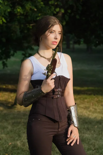 一个穿着紧身胸衣和裤子 带着板牙的年轻貌美的女人手里拿着一把双管火枪 一个非正式的蒸汽灌篮模型在一个夏季公园的夜晚 — 图库照片