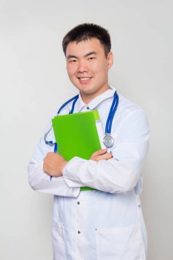 Boynunda steteskop olan beyaz önlüklü bir doktor elinde yeşil bir dosya tutuyor. Biri beyaz arkaplanda..