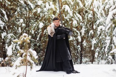 Siyah örtülü siyah pelerinli, beyaz kürklü ve elinde uzun kılıçlı bir savaşçı çam ormanının arka planında derin bir karda yürüyor..