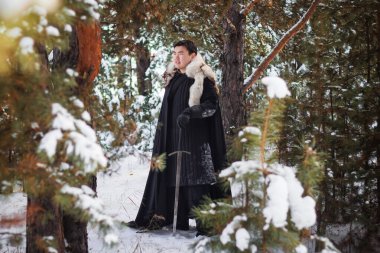 Siyah örtülü siyah pelerinli, beyaz kürklü ve elinde uzun kılıçlı bir savaşçı çam ormanının arka planında derin bir karda yürüyor..