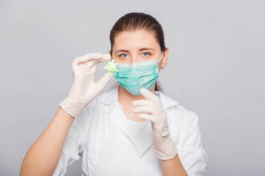 Beyaz önlüklü, maskeli ve eldivenli doktor elinde sahte bir virüs tutuyor. Beyaz arkaplanda stüdyo fotoğrafı.