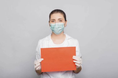 Beyaz önlüklü, maskeli ve eldivenli kadın doktor elinde kırmızı bir kağıt tutuyordu. Beyaz arkaplanda stüdyo fotoğrafı.