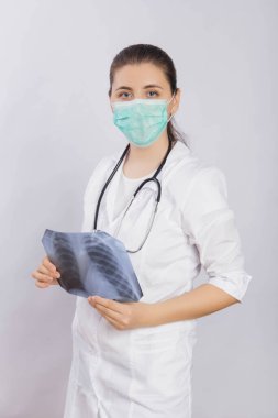 Beyaz önlüklü, maskeli ve boynunda steteskop olan çekici bir kadın doktor ellerinde bir röntgen tutuyor..