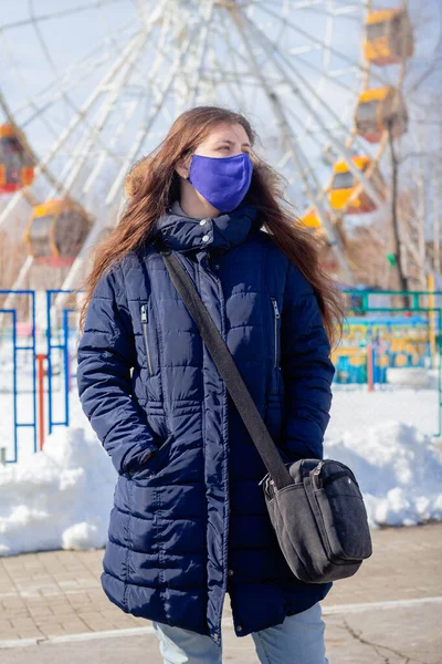 Portrait of a girl in a winter jacket with a hood and in a blue protective mask on her face strolling through a deserted amusement park.