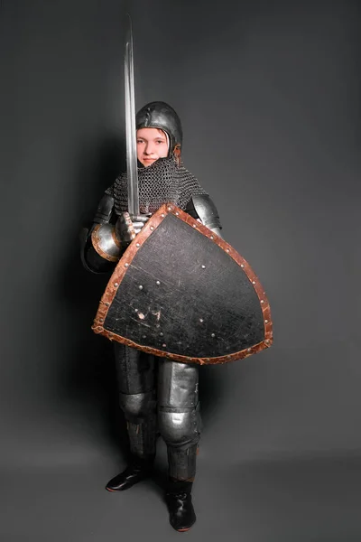 A young knight in medieval armor armed with a sword and shield standing on a gray background.