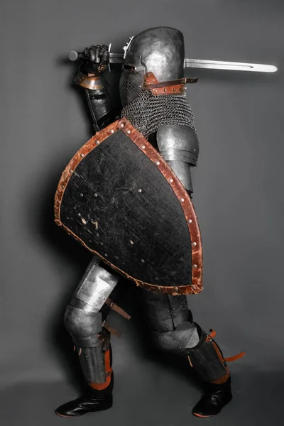 A medieval knight in armor with a shield and a sword in his hands stands in a fighting position and waves his weapon.