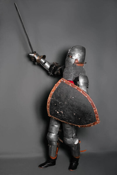 A medieval knight in armor with a shield and a sword in his hands stands in a fighting position and waves his weapon.