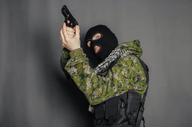 Kamuflaj üniformalı, çelik yelekli ve kar maskeli bir adam silahını hazır tutuyor ve gri arka planda duran bir tabancayla nişan alıyor..