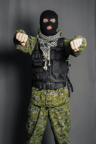 A man in camouflage and bullet proof vest, with a balaclava on his head. A special unit soldier, a modern warrior. A photo on a gray background, studio.