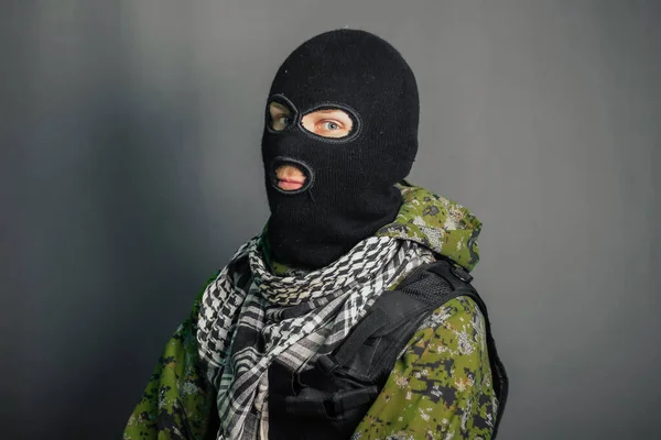 A man in camouflage and bullet proof vest, with a balaclava on his head. A special unit soldier, a modern warrior. A photo on a gray background, studio.