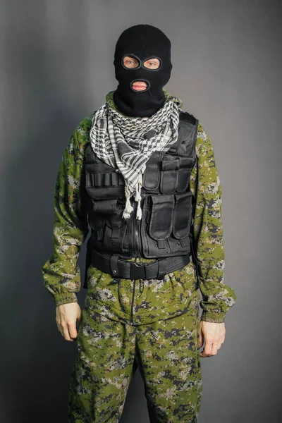 A man in camouflage and bullet proof vest, with a balaclava on his head. A special unit soldier, a modern warrior. A photo on a gray background, studio.