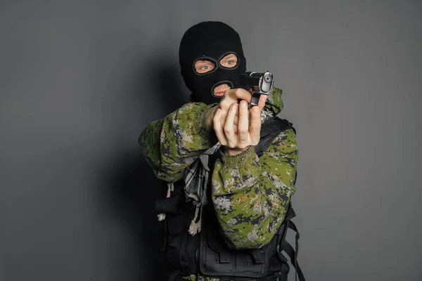 A man in camouflage uniform, body armor and a balaclava, holds his weapon ready and takes aim with a pistol, standing against a gray background.