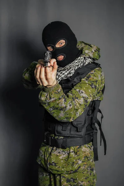 A man in camouflage uniform, body armor and a balaclava, holds his weapon ready and takes aim with a pistol, standing against a gray background.