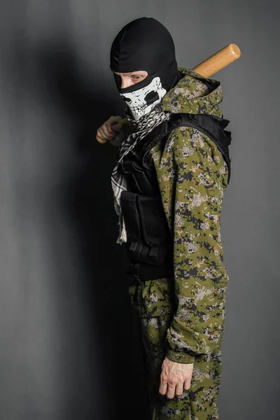 Unknown man, a robber in a balaclava with a skull, in camouflage uniform and body armor with a baseball bat in his hands. Studio photo on a gray background.