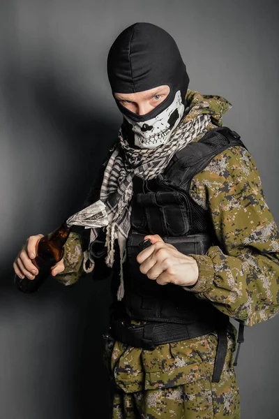 A man in a balaclava with a skull pattern, dressed in camouflage with a hood and a bulletproof vest, is holding a Molotov cocktail in his hands. A protester throws a Molotov cocktail. Studio photo on a gray background.