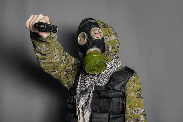 A soldier in camouflage uniform, body armor, in a gas mask with a hood on his head, holds a gun in his hands. Cosplay man, stalker. Studio photo on a gray background.