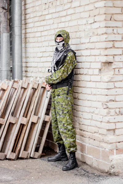 A man in camouflage, balaclava with a skull and bulletproof vest, with a baseball bat in his hands, against the background of a brick wall. Gangster is preparing an attack, from the corner.