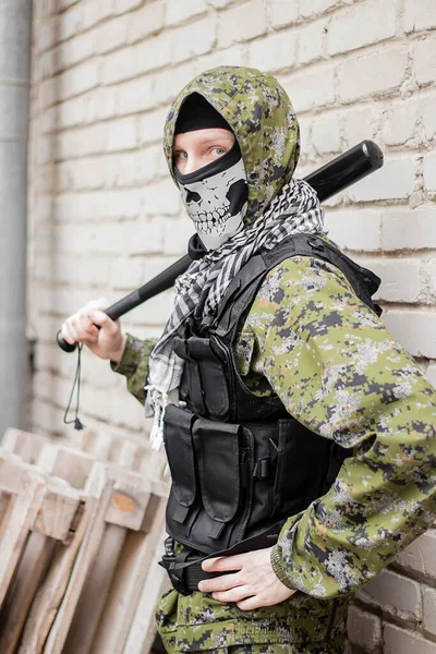 A man in camouflage, balaclava with a skull and bulletproof vest, with a baseball bat in his hands, against the background of a brick wall. Gangster is preparing an attack, from the corner.